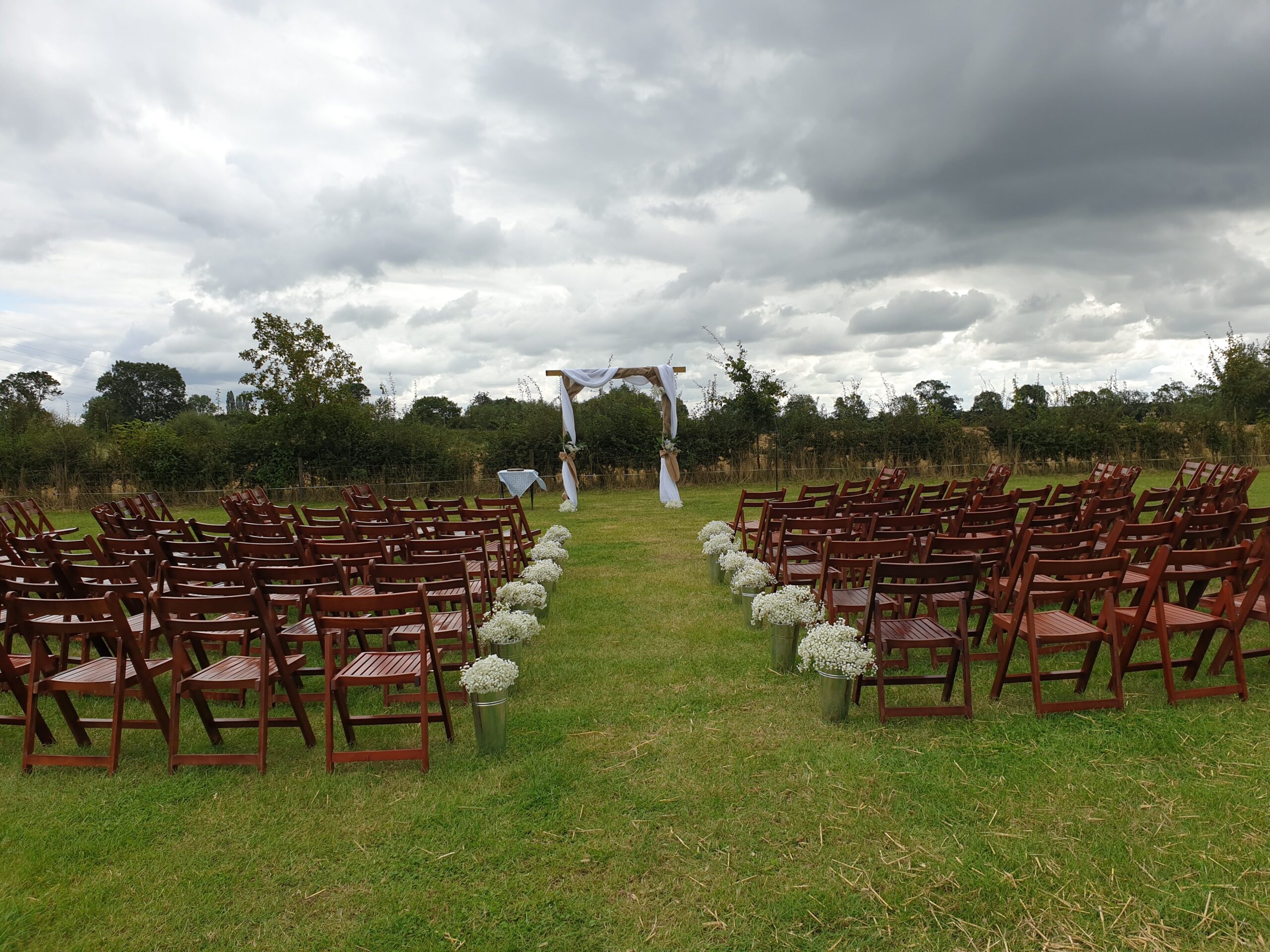 A field is set up with rows or dark oak rustic chairs facing an arch and lined with tin containers