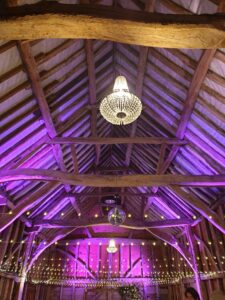 purple disco lights shine onto the beamed ceiling of a barn with a chandlier