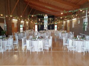 village hall with wooden floor and woodne clad ceiling, white clothed tables and chivari chairs and lights strung across the room