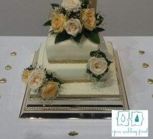 a two tier square cake with cream and ivory flowers on a square rose gold metal cake stand 