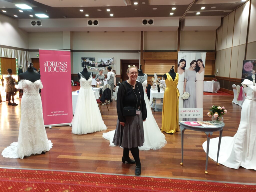 Your Wedding Friend is stood in front of a selection of wedding dresses at a wedding fair