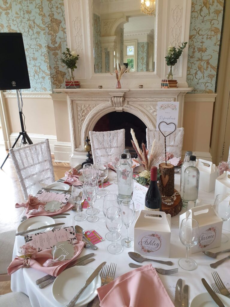 weddin breakfast table set with a rustic log centre piece and baby pink napkins folded onto a plate, in front of an elegant white fireplace with mirror