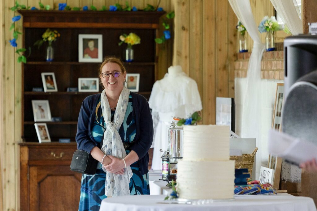 Your wedding friend is stood in front of a dresser, next to a wedding cake at a barn venue introducing the speeches